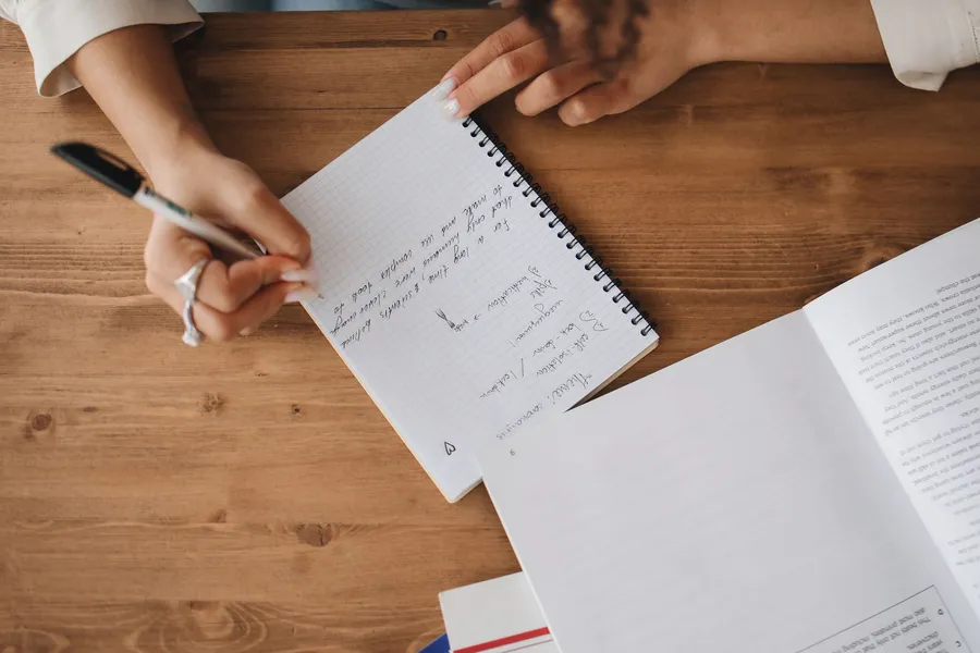A person writing a list of questions in a notebook at a desk
